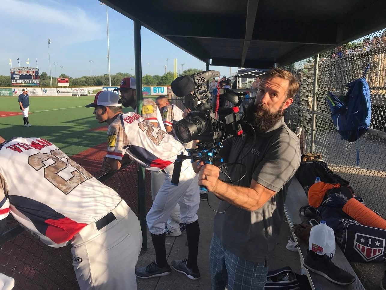 A cameraman films a baseball game from the dugout as players watch the field.