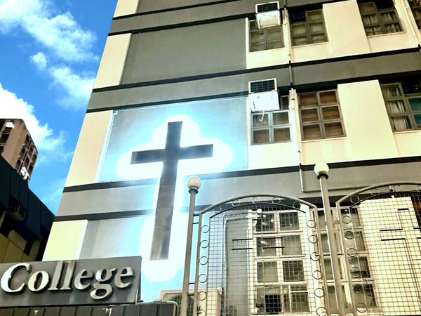 A bright cross glows on a college building wall under a blue sky.