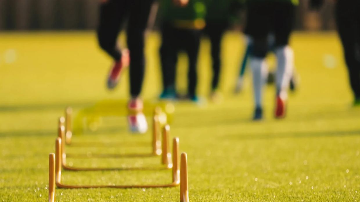 Athletes running agility drills over hurdles on a grassy field.