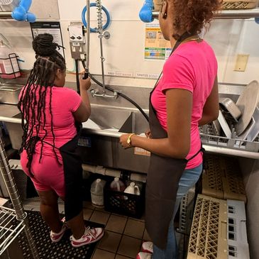 Two women in pink shirts washing dishes in a kitchen sink area.