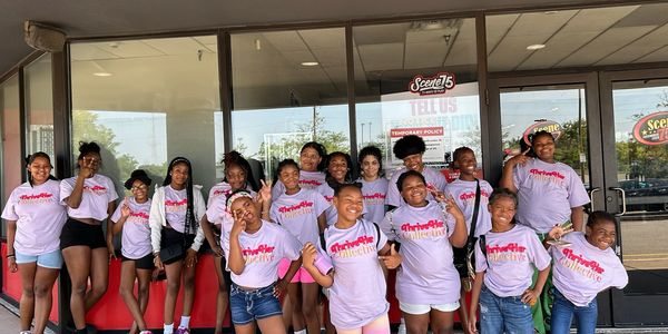 Group of smiling girls wearing matching ThriveHer Collective t-shirts outside a storefront.