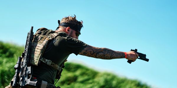 Tattooed soldier aiming and firing a handgun outdoors.