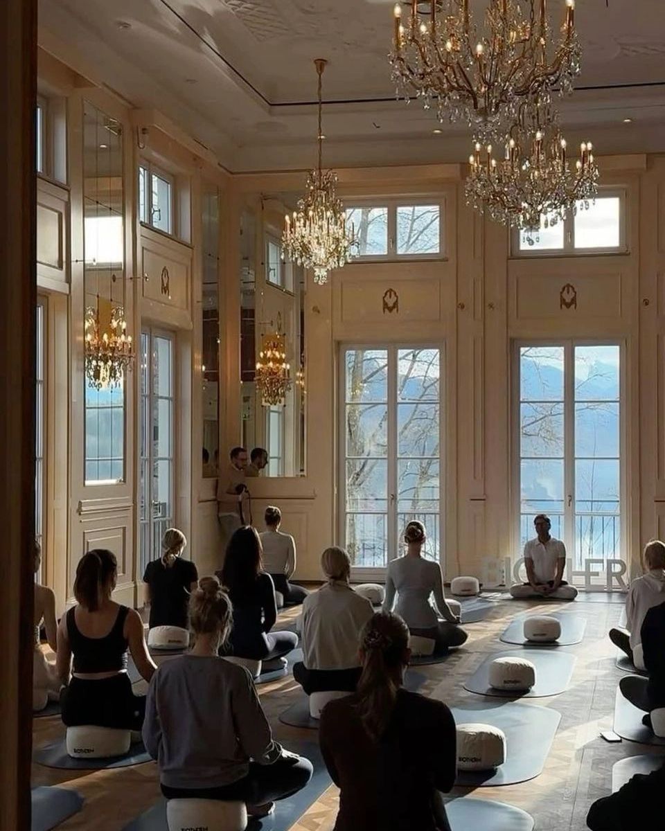 Group meditation session in an elegant, chandelier-lit room with large windows overlooking nature.