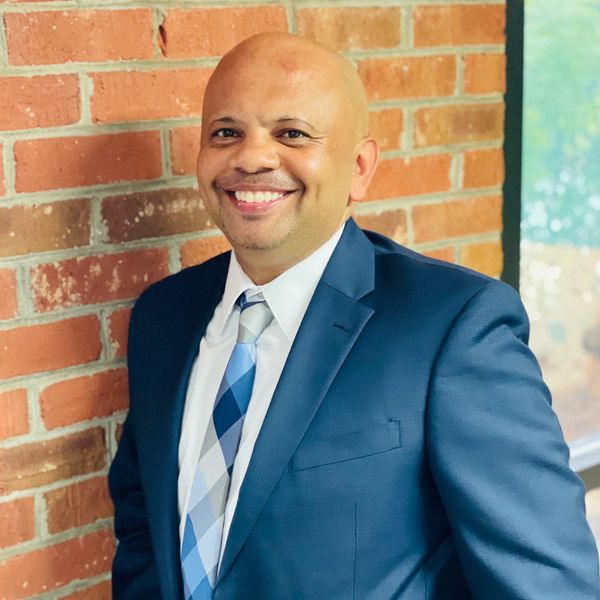 Smiling man in a blue suit and patterned tie leaning against a brick wall.