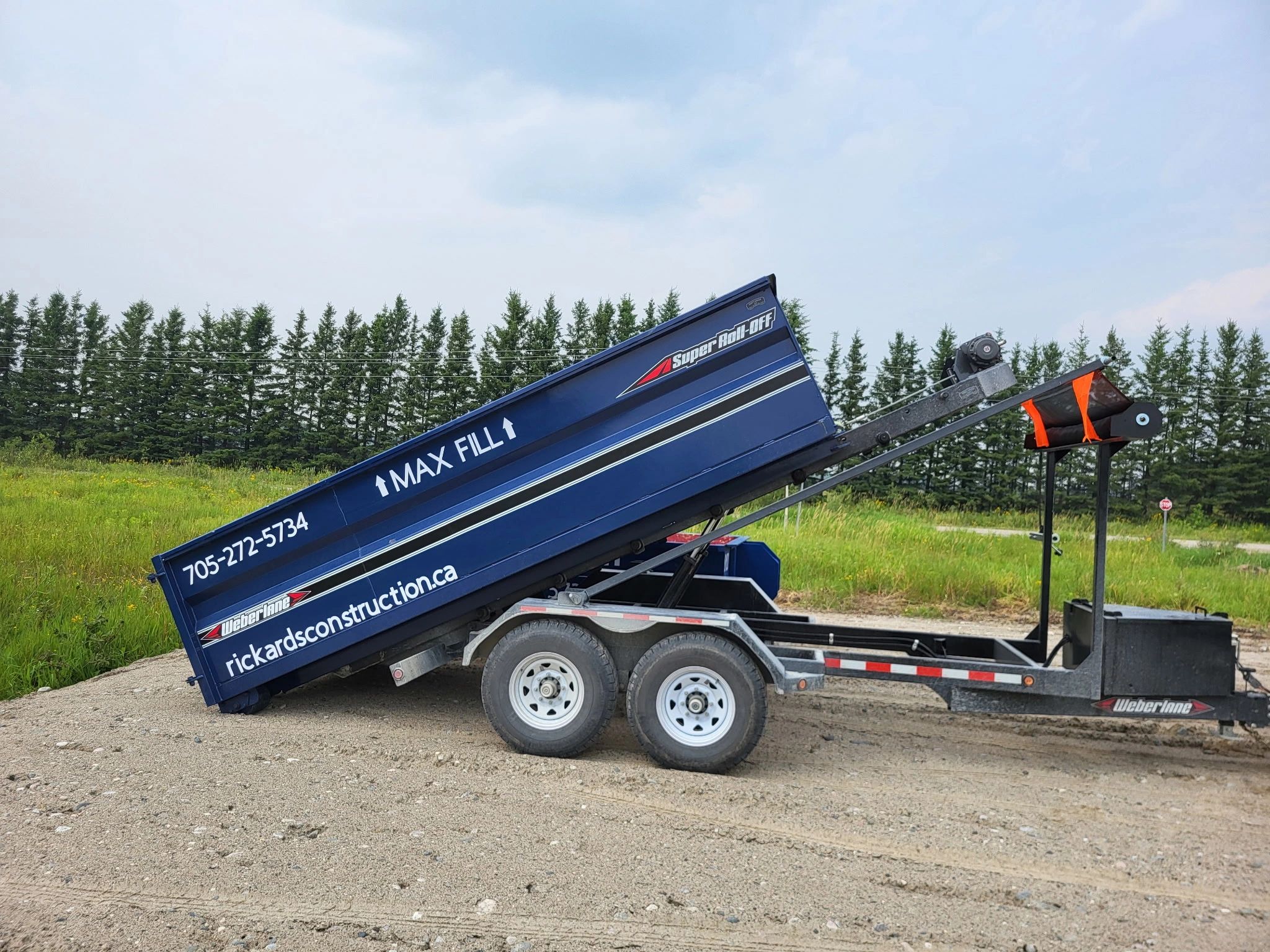 Blue construction trailer with raised bed on gravel near trees.