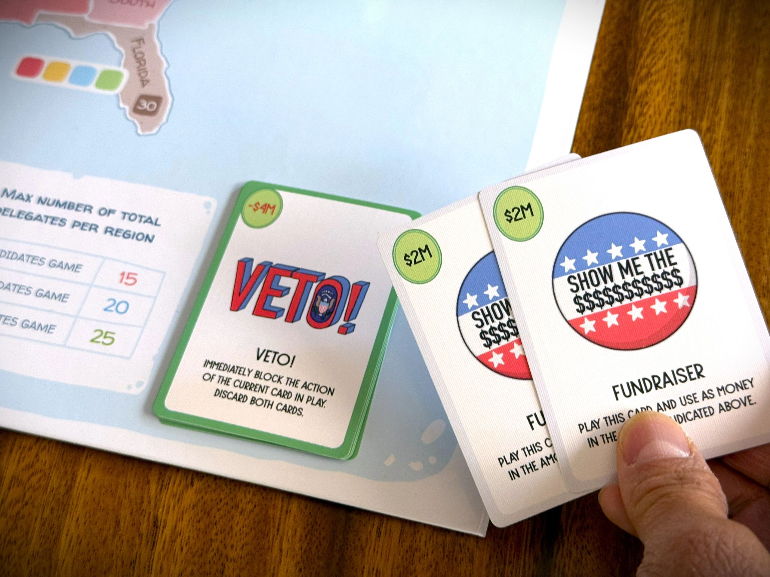 Hand holding political game cards: Veto and Fundraiser cards with a game map in background.