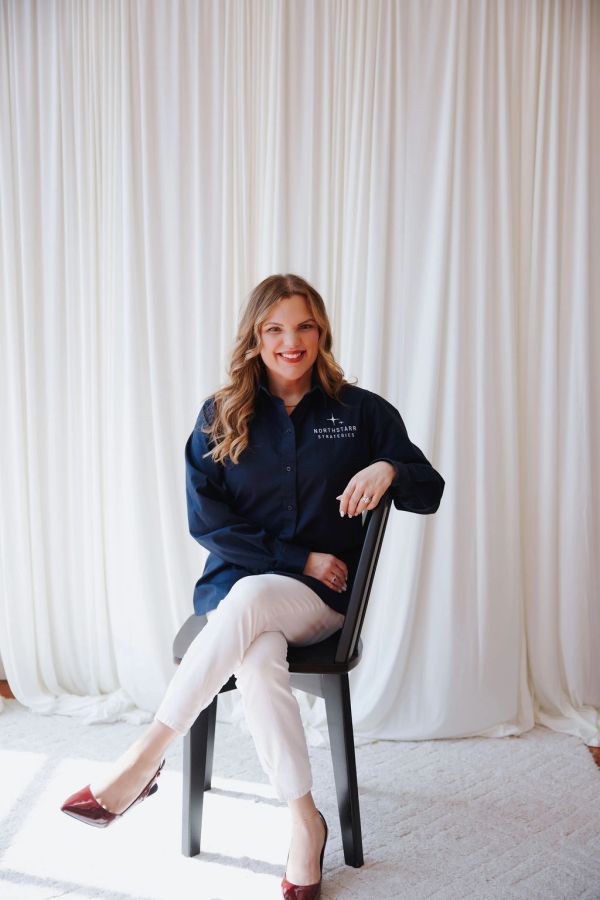 Woman sitting on chair smiling, wearing a navy shirt and white pants.