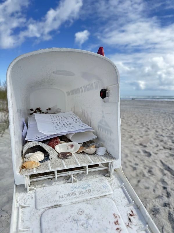 Open mailbox with shells and papers on a sandy beach.