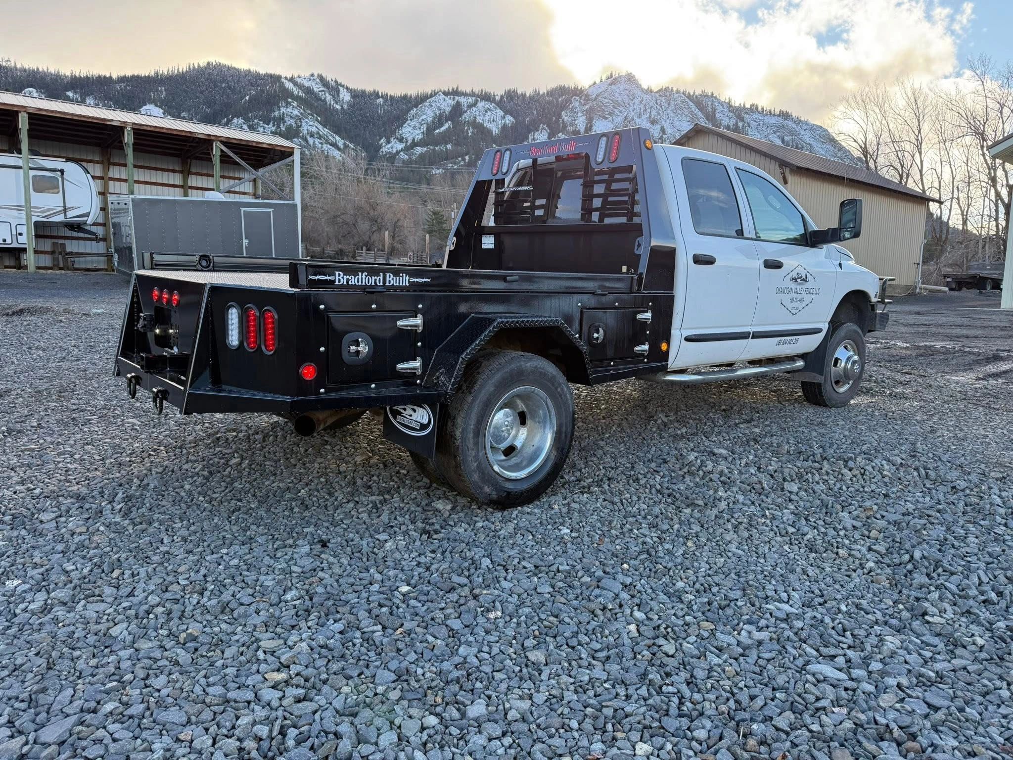 White truck with black flatbed parked on gravel with snowy mountains in background.