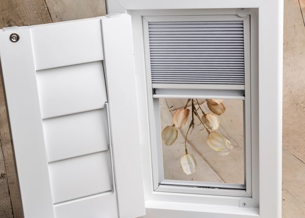 White window frame with closed shutter and flowers reflected in the glass.
