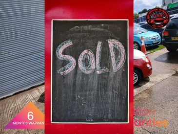 Red and white compact car parked outside a garage with warranty logos.