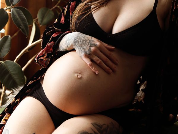 Pregnant woman in black lingerie with tattoos gently holding her belly.