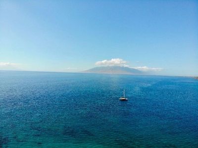 A sailboat floats on calm blue waters with a mountainous island in the distance under a clear sky.