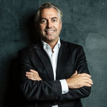 Confident man in a black suit smiling with arms crossed against a dark textured wall.