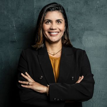 Confident woman in black blazer smiling with arms crossed against a dark textured wall.