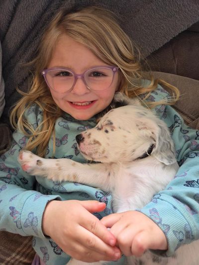 A smiling girl with glasses cuddles a sleepy spotted puppy.