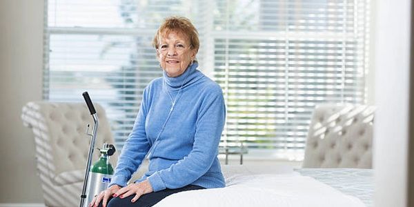 Lady sitting on bed smiling while using a portable oxygen tank.