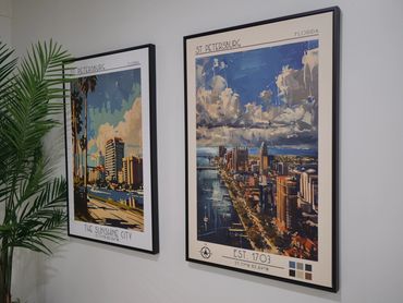 Two framed cityscape posters hang on a white wall beside a green plant.