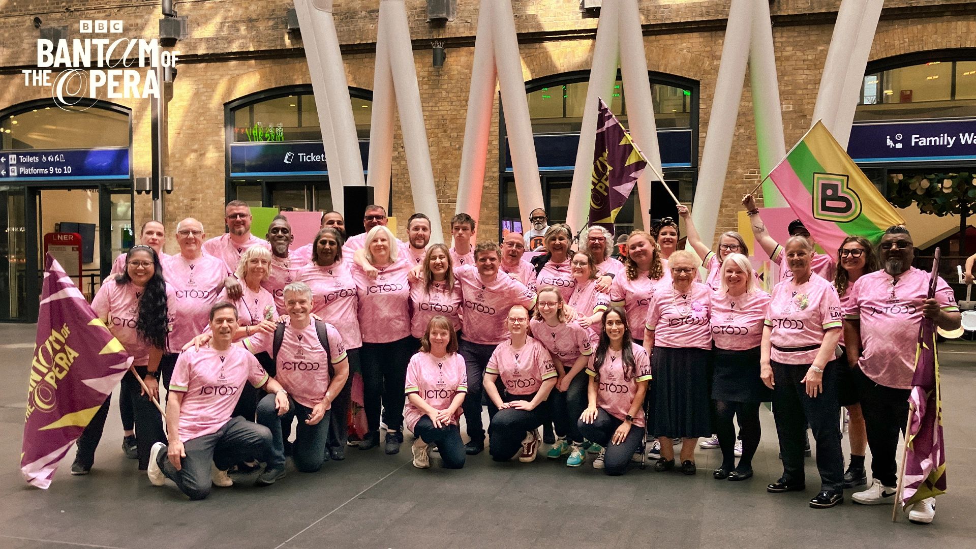 Group of people in pink shirts posing with flags at a train station event.