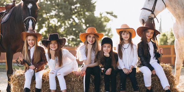 Six girls in cowboy hats sitting on hay bales with horses behind them.