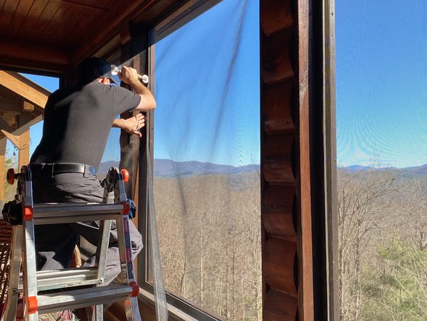 Man installing a screen on a large window in a wooden cabin.