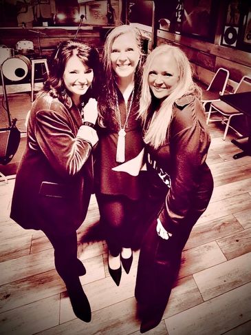 Three women smiling together in a warmly lit room with wooden floors.