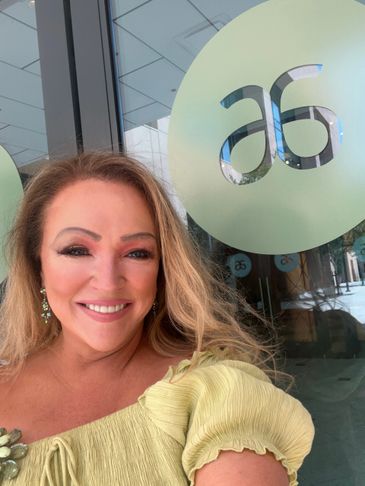 Smiling woman in yellow top taking a selfie near a glass door with 'as' logo.