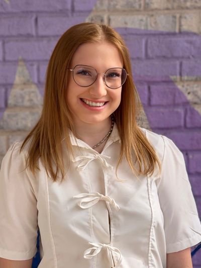 Young woman with glasses smiling in front of a purple brick wall.