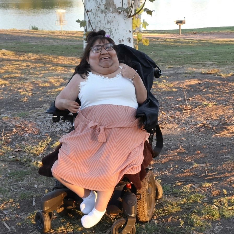 Woman in wheelchair smiling outdoors near a lake on a sunny day.