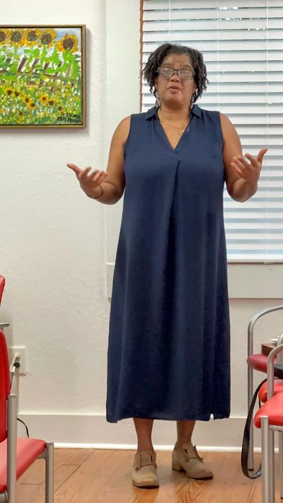 Woman in blue dress speaking in a room with red chairs and sunflower painting.