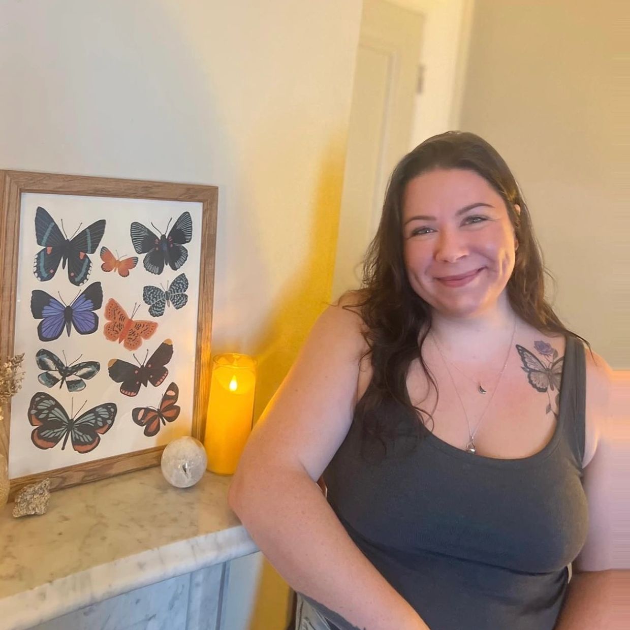 Smiling woman with butterfly tattoo next to butterfly artwork and candle.