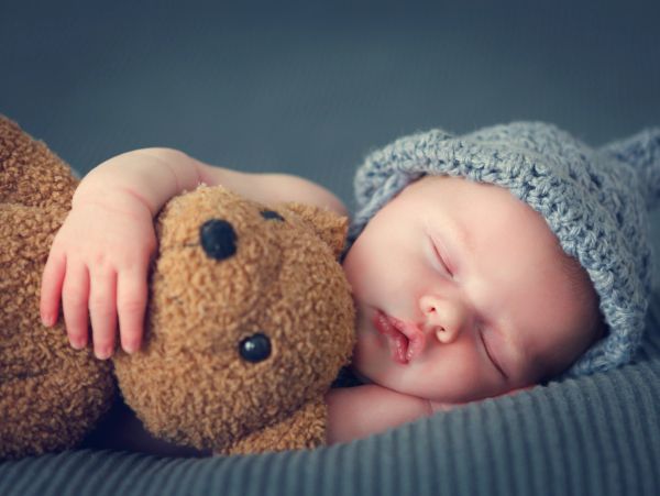 Sleeping baby cuddling a teddy bear, wearing a knitted hat.