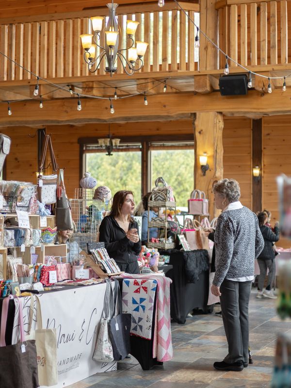 Two women talking at a handmade crafts market in a wooden indoor venue.