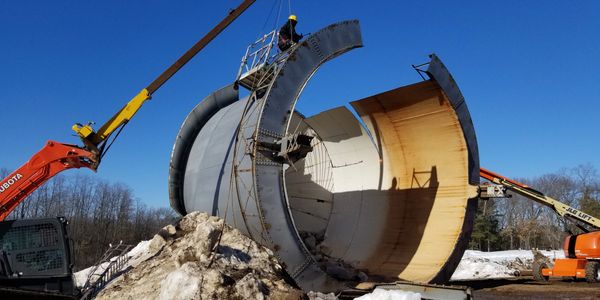 Workers use cranes to dismantle a large cylindrical metal structure outdoors.
