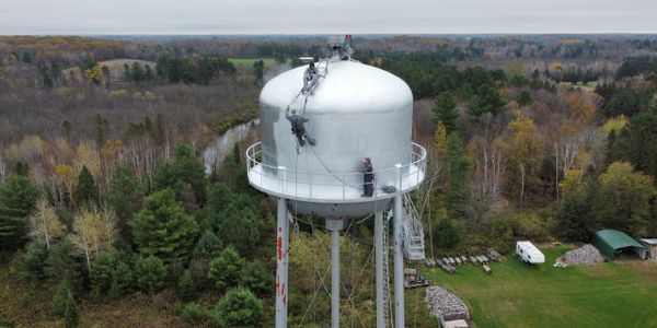 Workers maintain a large water tower surrounded by forest and fields.