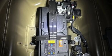 Car battery installed in vehicle trunk with wiring and straps.
