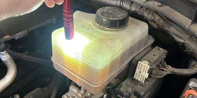A person uses a flashlight to inspect a translucent brake fluid reservoir in a car engine bay.