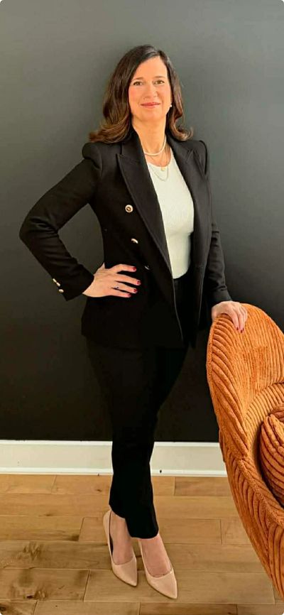 A confident woman in a black suit and white top stands beside an orange chair against a black wall.