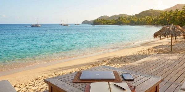Beachfront workspace with laptop, notebook, and phone overlooking ocean and boats.