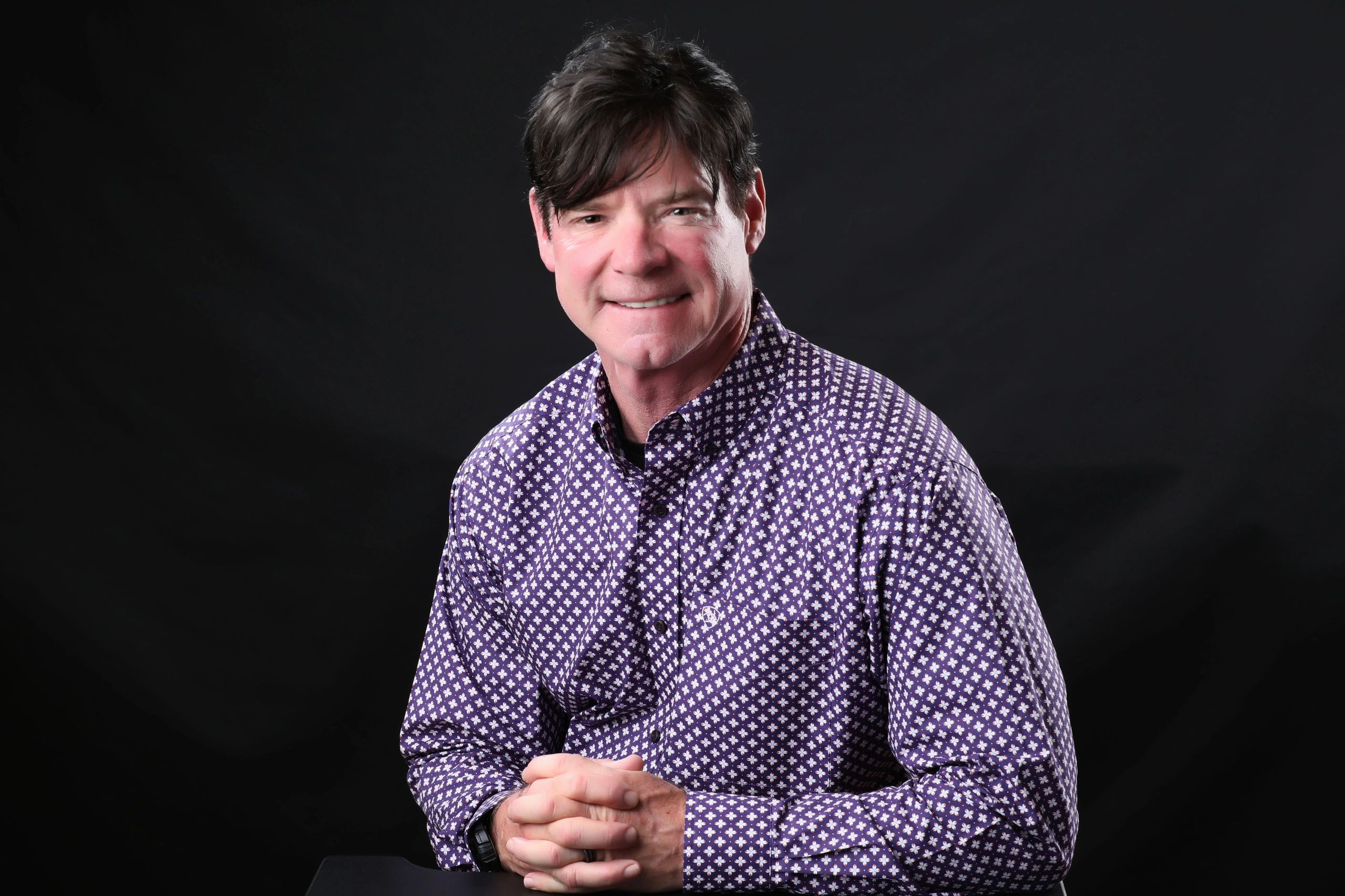 Smiling man in a patterned purple shirt against a black background.
