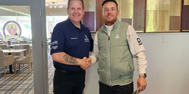 Two men shaking hands indoors, one in a blue shirt and the other in a light green vest.