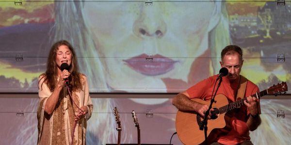 A woman sings while a man plays guitar on stage with a large face projection behind them.