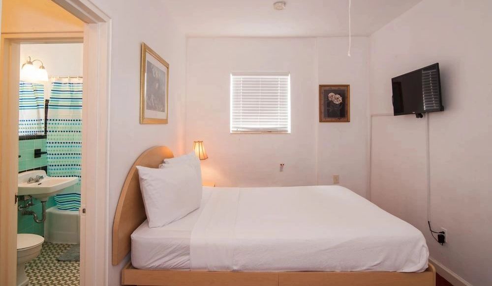 A small guest room with a bed, TV, and window, with an open door to a bathroom
