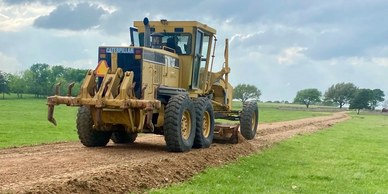A Caterpillar grader leveling a dirt path in a grassy field.