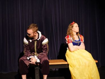 Two actors in costume sitting back-to-back on a bench on stage.