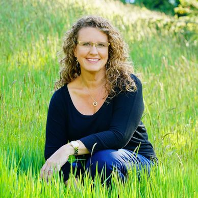 Woman with curly blonde hair and a smile sitting in tall green grass on a hill.