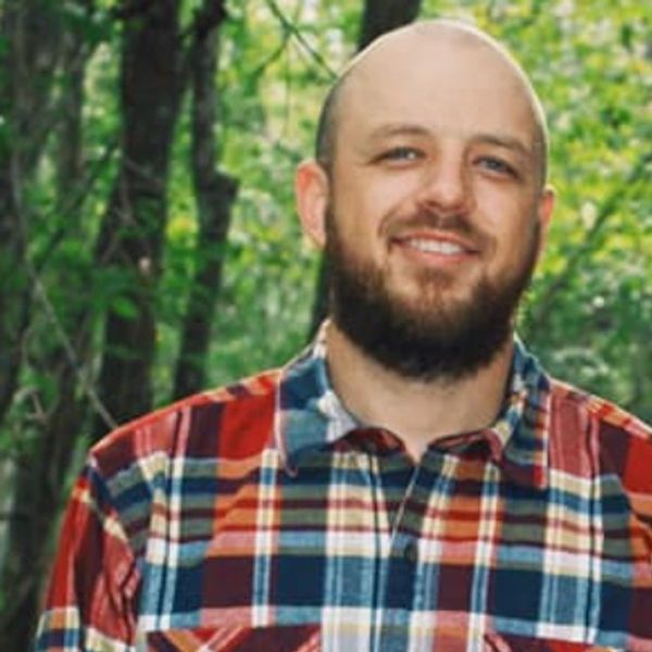 Smiling man with a beard wearing a colorful plaid shirt in a forest.