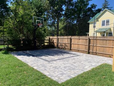 A backyard basketball court with a hoop and paved surface surrounded by grass and a wooden fence.