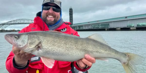 Man in red jacket holding a large fish on a boat with a bridge in the background.