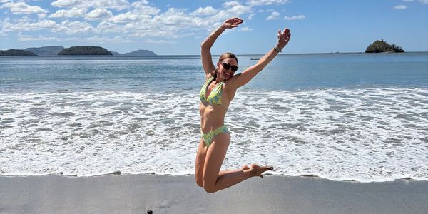 Woman in a green bikini joyfully jumping on a sunny beach with ocean waves behind her.